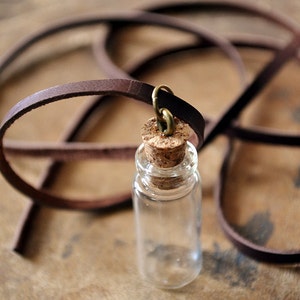 May include: A small glass bottle with a cork stopper and a brown leather cord attached. The bottle is empty and has a clear glass body.