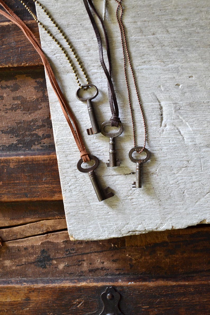 May include: Four antique skeleton keys on different chains, including a gold chain, a brown leather cord, and a black leather cord. The keys are displayed on a white wooden surface.