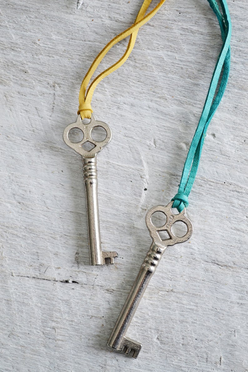 Op de afbeelding: Twee antieke skeletsleutels met sierlijke sleutelgaten, elk bevestigd aan een leren koord. Een koord is geel en de andere is turquoise.