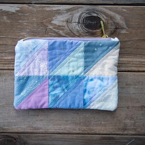 May include: A rectangular quilted zipper pouch featuring a patchwork design of various blue, purple, and white fabric triangles. The pouch has a gold-toned metal zipper closure along the top edge and displays a soft, stitched texture throughout.