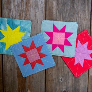 May include: Four colorful, square fabric coasters with starburst designs. The coasters feature a variety of colors, including yellow, red, pink, and blue, with quilted stitching. They are arranged on a rustic wooden surface.