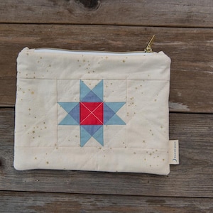 May include: Cream-colored quilted pouch with a star design. The star is composed of blue and red geometric shapes. The pouch has a gold zipper and a small tag on the side. The background is a rustic wooden surface.