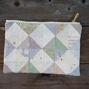 May include: A quilted pouch with a geometric pattern in shades of white, blue, and gold. The pouch has a gold zipper and is made of fabric squares sewn together. The design is reminiscent of a patchwork quilt.