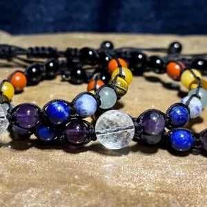 May include: Three beaded bracelets with a variety of colorful gemstones and black beads. The bracelets are made with a macrame knotting technique. The gemstones include clear quartz, amethyst, lapis lazuli, and tiger's eye.