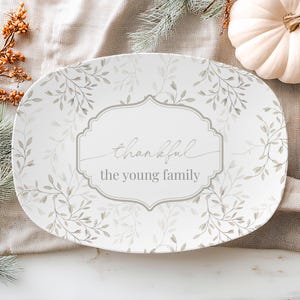 May include: An oval serving platter with a white background and a delicate leaf pattern in shades of gray and green. The center features the words "thankful" and "the young family" in elegant script. A white pumpkin and fall foliage are in the background.
