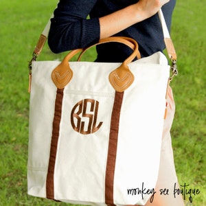 May include: A white canvas tote bag with brown leather accents and a brown monogrammed initial "BSL". The bag has a long strap and is being held by a person.