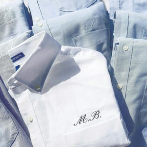 May include: A collection of button-down shirts in various shades of blue and white. Several shirts are embroidered with initials, and one has the text "AUTHORITY" on the collar. The shirts are neatly folded, showcasing their crisp collars and button details.