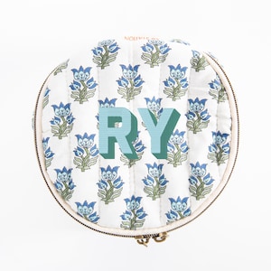 May include: Round, quilted cosmetic bag with a white background and a repeating blue floral pattern. The letters "RY" are in the center in a teal and green 3D font. The bag has a beige zipper and trim.