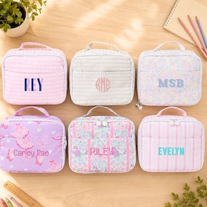 May include: Six rectangular zippered lunch bags with top handles, each featuring unique patterns including pink gingham, beige gingham, floral prints, ballet motifs, and pink stripes. Each bag is personalized with embroidered names or initials.