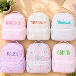 May include: Six small backpacks with top handles and front zippered pockets. The bags feature various patterns including pink gingham, beige gingham, floral prints, and stripes. Each backpack is personalized with embroidered names: Mackenzie, Anna Grace, Madison, Riley, MSB, and Ashlyn.