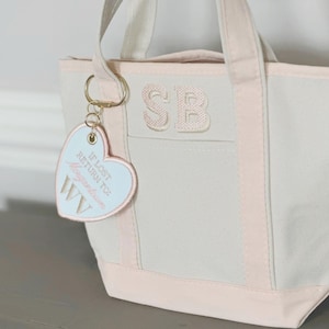May include: A canvas tote bag with light pink trim and handles. The bag features the embroidered initials "SB" and a heart-shaped keychain with the text "IF LOST RETURN TO: Morgantown WV".