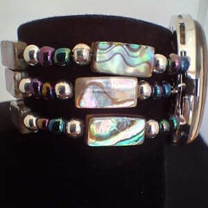 Pu&ograve; includere: Un bracciale a pi&ugrave; fili con tre file di perline rettangolari in conchiglia di abalone dai toni iridescenti verdi, rosa e blu. Le file sono arricchite da perline sferiche color argento metallizzato e viola scuro, collegate a una cassa di orologio argentata.