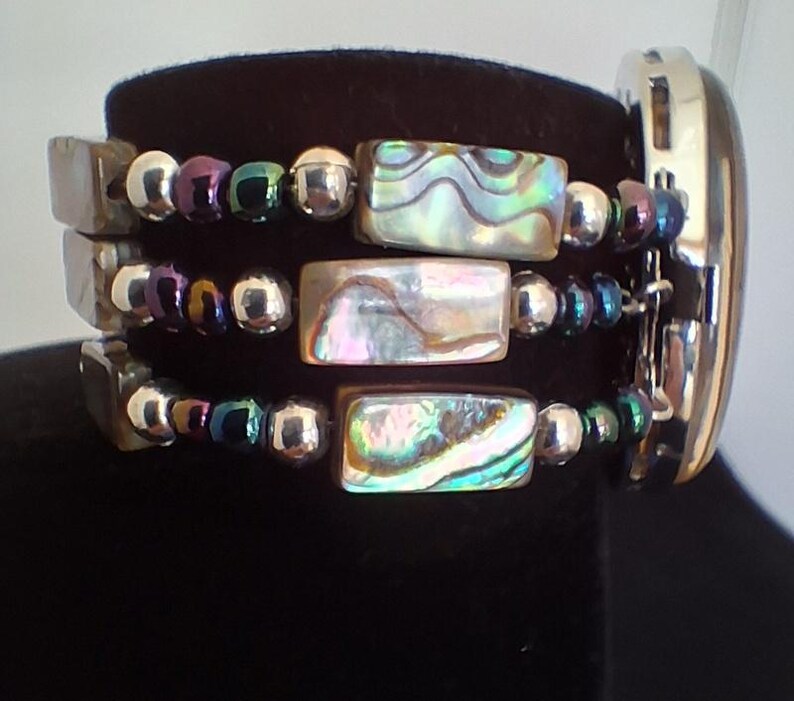 Pu&ograve; includere: Un bracciale a pi&ugrave; fili con tre file di perline rettangolari in conchiglia di abalone dai toni iridescenti verdi, rosa e blu. Le file sono arricchite da perline sferiche color argento metallizzato e viola scuro, collegate a una cassa di orologio argentata.