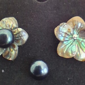 Pu&ograve; includere: Due componenti a forma di fiore intagliati in conchiglia di abalone iridescente, uno con una perla scura, rotonda e lucida al centro. Una singola perla scura, rotonda e lucida &egrave; posizionata sotto i fiori. La lettera B &egrave; visibile.