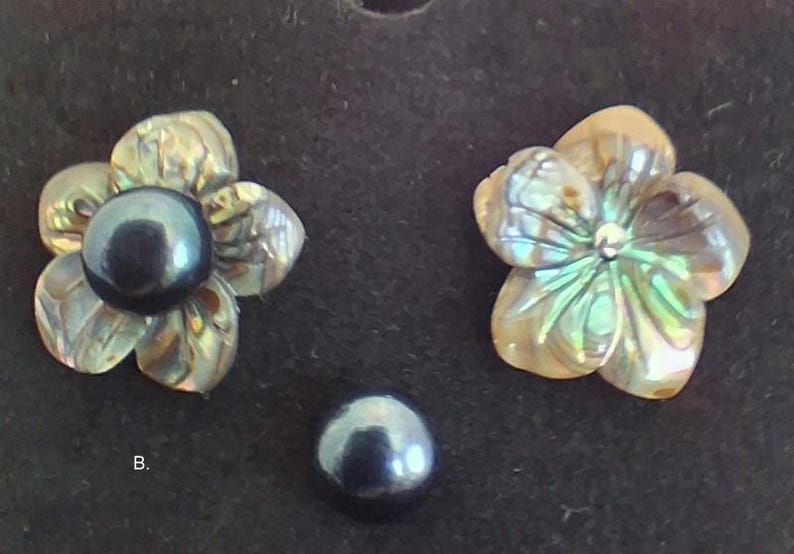 Pu&ograve; includere: Due componenti a forma di fiore intagliati in conchiglia di abalone iridescente, uno con una perla scura, rotonda e lucida al centro. Una singola perla scura, rotonda e lucida &egrave; posizionata sotto i fiori. La lettera B &egrave; visibile.