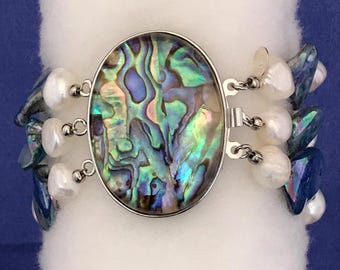 May include: A silver bracelet with a large abalone shell charm and white pearls. The bracelet is made of multiple strands of pearls and abalone shell pieces.