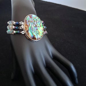 Pu&ograve; includere: Un bracciale a pi&ugrave; fili caratterizzato da un grande elemento centrale ovale in conchiglia di abalone con vortici iridescenti verdi, blu e marroni. Il bracciale include tre file di piccole perline quadrate di abalone alternate a perline metalliche scure.