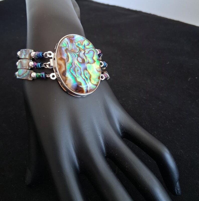 Pu&ograve; includere: Un bracciale a pi&ugrave; fili caratterizzato da un grande elemento centrale ovale in conchiglia di abalone con vortici iridescenti verdi, blu e marroni. Il bracciale include tre file di piccole perline quadrate di abalone alternate a perline metalliche scure.
