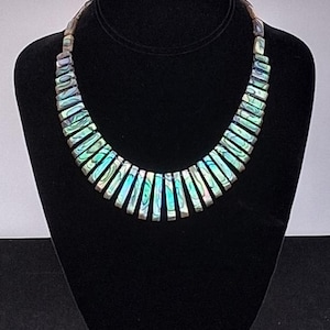 Pu&ograve; includere: Una collana caratterizzata da una serie di segmenti rettangolari in conchiglia di abalone disposti a ventaglio. Ogni segmento mostra sfumature iridescenti di verde, blu e viola, collegate per formare una forma a colletto curva.