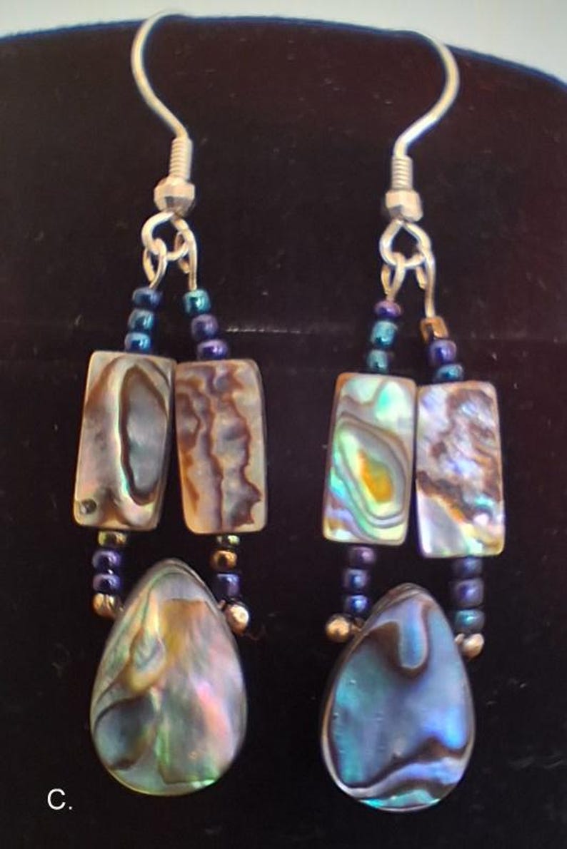 Pu&ograve; includere: Un paio di orecchini pendenti con elementi in conchiglia di abalone iridescente. Ogni orecchino presenta due pezzi rettangolari sospesi sopra un ciondolo a goccia, collegati da piccole perline blu scuro e metallizzate su ganci color argento.