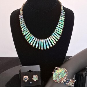 Pu&ograve; includere: Un set di gioielli composto da una collana con segmenti di conchiglia di abalone iridescente, un bracciale con un elemento centrale ovale in conchiglia di abalone e fili di perline, e orecchini a lobo a forma di fiore, etichettato Set B.