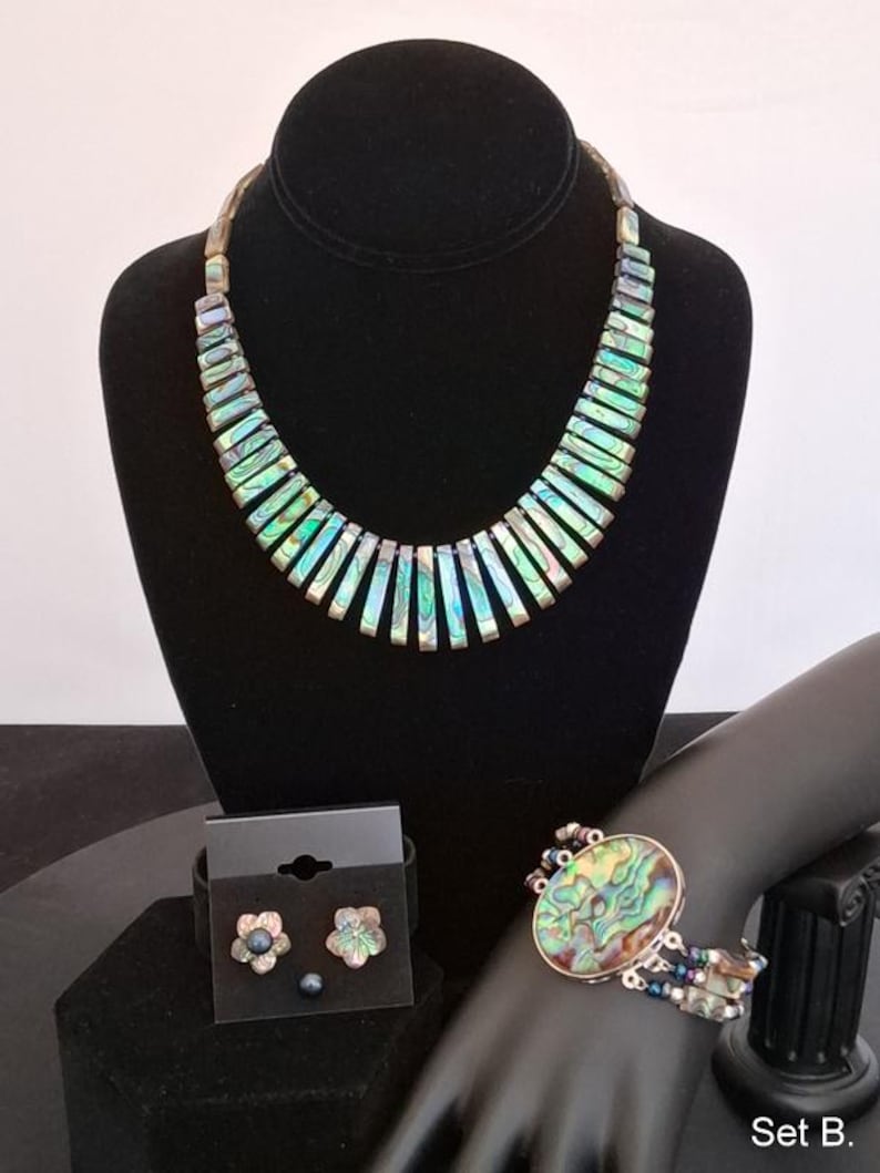 Pu&ograve; includere: Un set di gioielli composto da una collana con segmenti di conchiglia di abalone iridescente, un bracciale con un elemento centrale ovale in conchiglia di abalone e fili di perline, e orecchini a lobo a forma di fiore, etichettato Set B.