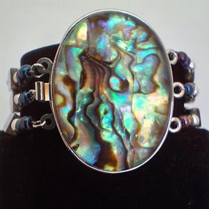 Pu&ograve; includere: Un bracciale con un grande elemento centrale ovale in conchiglia di abalone, caratterizzato da vortici iridescenti verdi, blu e marroni. L'elemento centrale &egrave; incastonato in una cornice argentata e collegato a tre fili di perline metalliche scure.