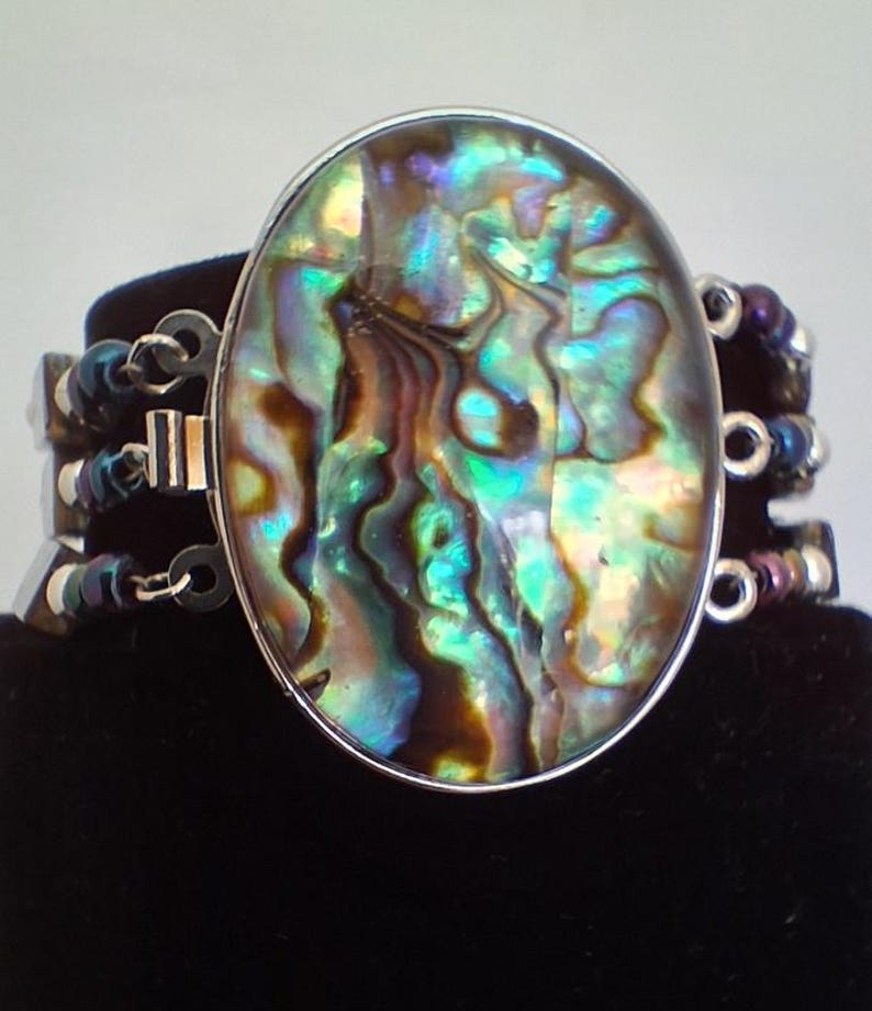 Pu&ograve; includere: Un bracciale con un grande elemento centrale ovale in conchiglia di abalone, caratterizzato da vortici iridescenti verdi, blu e marroni. L'elemento centrale &egrave; incastonato in una cornice argentata e collegato a tre fili di perline metalliche scure.