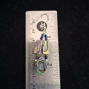 Pu&ograve; includere: Un orecchino pendente con due pezzi rettangolari di conchiglia di abalone e un pezzo a forma di goccia, arricchito da piccole perline blu iridescenti e un gancio color argento, mostrato accanto a un righello metallico di 3,8 cm.