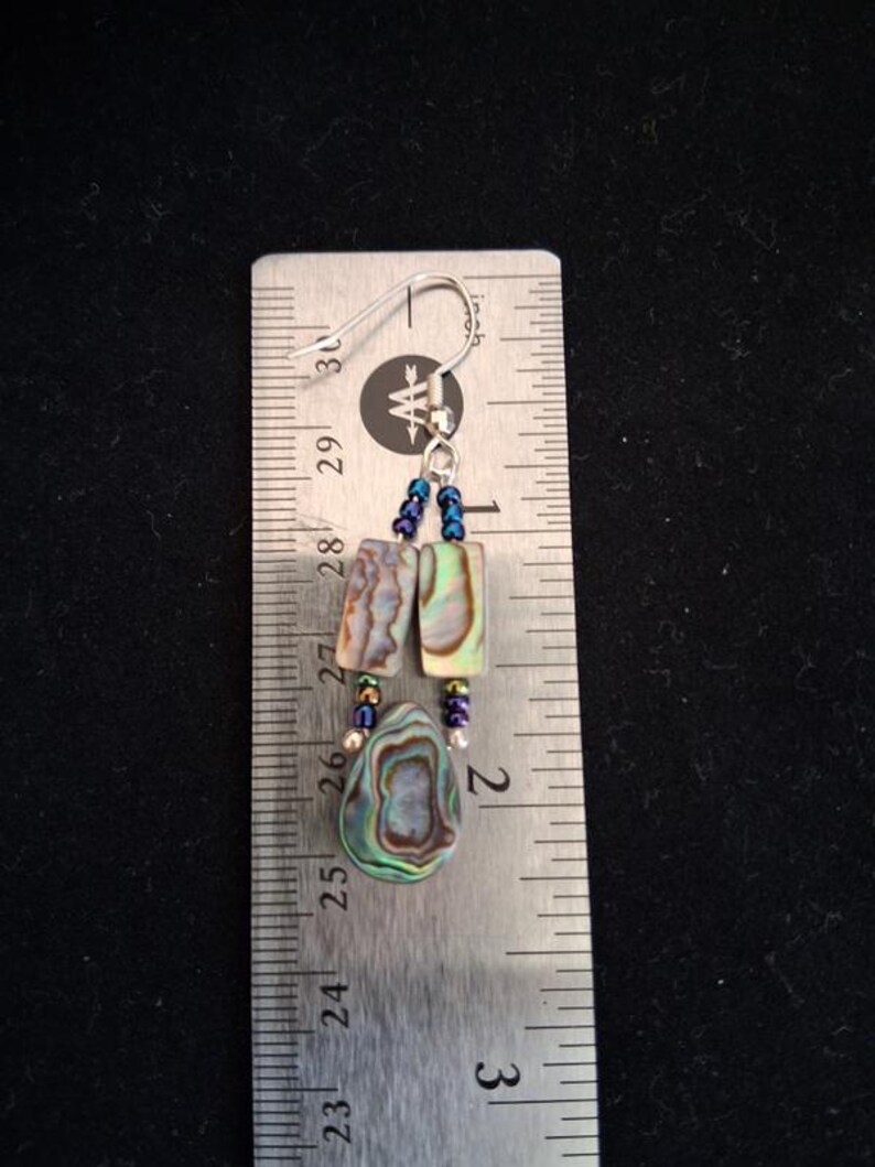 Pu&ograve; includere: Un orecchino pendente con due pezzi rettangolari di conchiglia di abalone e un pezzo a forma di goccia, arricchito da piccole perline blu iridescenti e un gancio color argento, mostrato accanto a un righello metallico di 3,8 cm.