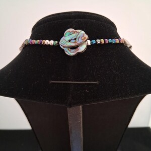 Pu&ograve; includere: Una collana con un bottone centrale a forma di fiore realizzato in conchiglia di abalone iridescente. Il filo &egrave; composto da piccole perline metalliche multicolori nei toni del blu, verde, viola e argento, disposte in un motivo ripetitivo.