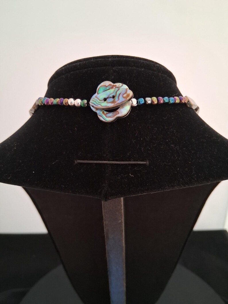 Pu&ograve; includere: Una collana con un bottone centrale a forma di fiore realizzato in conchiglia di abalone iridescente. Il filo &egrave; composto da piccole perline metalliche multicolori nei toni del blu, verde, viola e argento, disposte in un motivo ripetitivo.