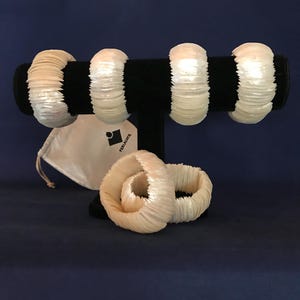 Puede incluir: Conjunto de seis pulseras de concha blanca en un soporte negro. Las pulseras están hechas de pequeñas conchas superpuestas. Hay una bolsa de cordón blanco con la palabra "remarkable" impresa en ella.