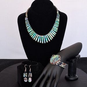 Pu&ograve; includere: Un set di gioielli etichettato Set C. include una collana composta da segmenti rettangolari di conchiglia di abalone iridescente, un bracciale con un grande elemento centrale ovale in abalone e dettagli a pi&ugrave; fili, e orecchini pendenti con perline.