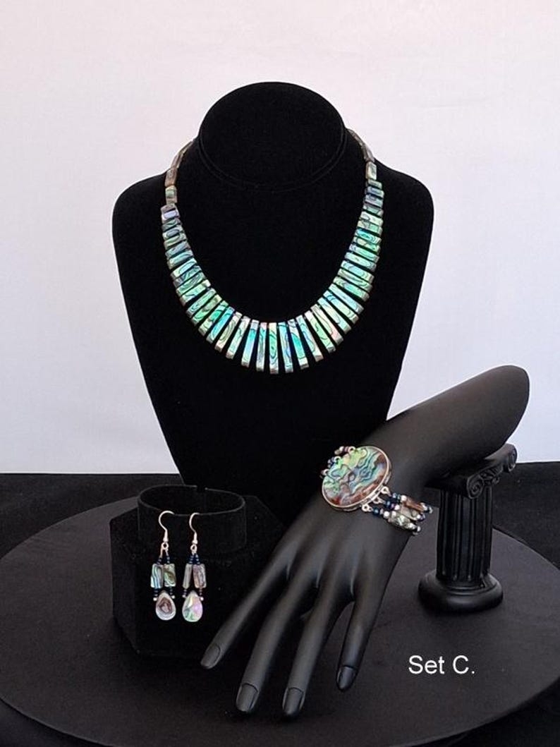 Pu&ograve; includere: Un set di gioielli etichettato Set C. include una collana composta da segmenti rettangolari di conchiglia di abalone iridescente, un bracciale con un grande elemento centrale ovale in abalone e dettagli a pi&ugrave; fili, e orecchini pendenti con perline.