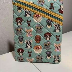 May include: A quilted teal fabric case featuring a repeating pattern of various dog breeds wearing sunglasses and accessories. The case has a gold zipper and a diamond-stitched pattern.