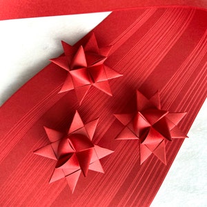 May include: Three red origami stars made from folded paper strips. The stars are arranged on a red paper background.