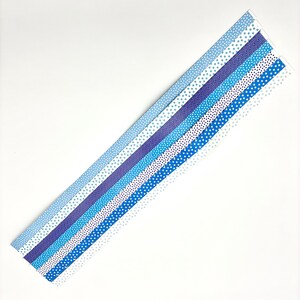 May include: An assortment of blue patterned paper strips, each with a unique design. The patterns include stars and dots in varying shades of blue, from light to dark. The strips are arranged diagonally against a white background.