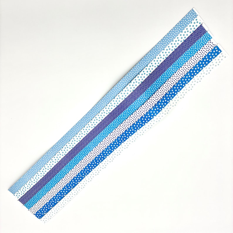 May include: An assortment of blue patterned paper strips, each with a unique design. The patterns include stars and dots in varying shades of blue, from light to dark. The strips are arranged diagonally against a white background.