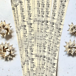 May include: Four star-shaped paper ornaments and several strips of sheet music are arranged on a light gray surface. The ornaments are made from folded sheet music, displaying black musical notes on a cream background. The sheet music strips are also cream with black notes.