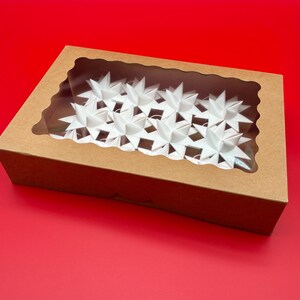 Może przedstawiać: Brązowe kartonowe pudełko z przezroczystym okienkiem, wypełnione białymi dekoracjami w kształcie gwiazd. Pudełko stoi na czerwonej powierzchni, a gwiazdy są ułożone w rzędach. Okienko pozwala na widok zawartości.