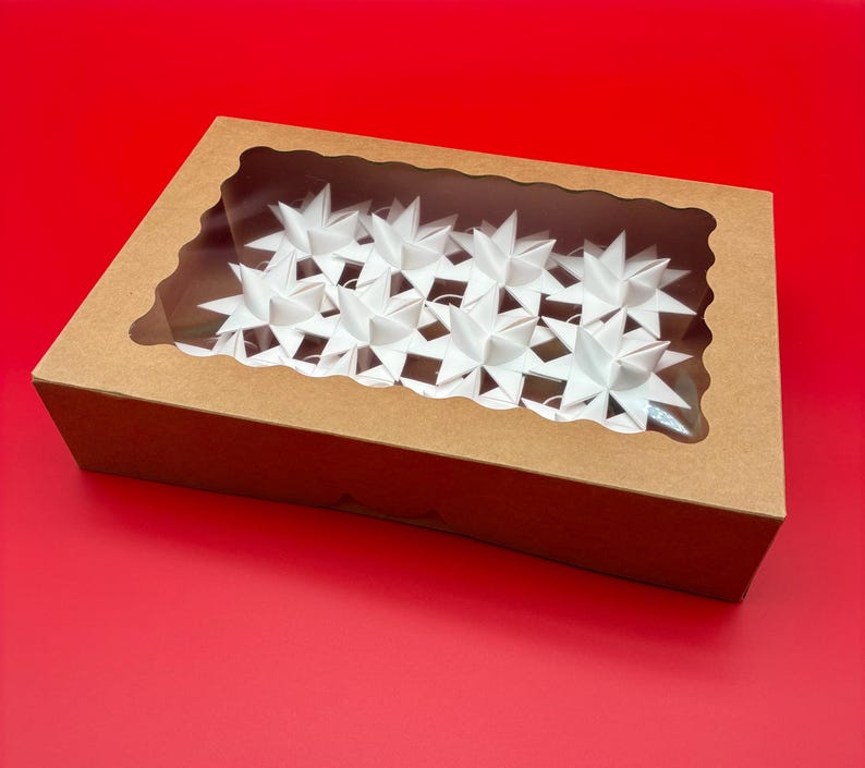 Może przedstawiać: Brązowe kartonowe pudełko z przezroczystym okienkiem, wypełnione białymi dekoracjami w kształcie gwiazd. Pudełko stoi na czerwonej powierzchni, a gwiazdy są ułożone w rzędach. Okienko pozwala na widok zawartości.