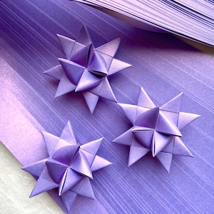 May include: Three purple origami stars on a purple background with a white border. The stars are made of folded paper and have a three-dimensional appearance.