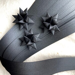 May include: Three black paper stars, folded into a 3D shape, are arranged on a black paper ribbon. The ribbon is folded in a wave pattern.