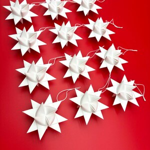Może przedstawiać: Szereg białych papierowych ozd&oacute;b w kształcie gwiazd na żywym czerwonym tle. Każda gwiazda ma wiele punkt&oacute;w i jest połączona cienkim białym sznurkiem. Gwiazdy są r&oacute;wnomiernie rozmieszczone, tworząc świąteczną i dekoracyjną ekspozycję.