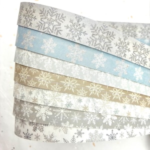 May include: A stack of decorative paper ribbons in various colors, each adorned with a snowflake pattern. The ribbons include shades of silver, blue, beige, and gray, creating a winter or holiday theme.