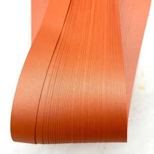 May include: Close-up of a stack of terracotta-colored paper strips. The strips are tightly packed together, with visible vertical lines. The paper has a slightly textured surface and a warm, earthy tone. The edges of the paper are curved.