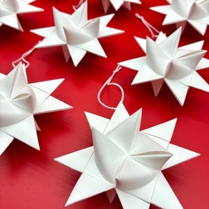 Może przedstawiać: Białe papierowe ozdoby w kształcie gwiazd z ostrymi krawędziami, ułożone na czerwonym tle. Każda gwiazda ma sznurek do zawieszenia. Gwiazdy to świąteczna dekoracja.