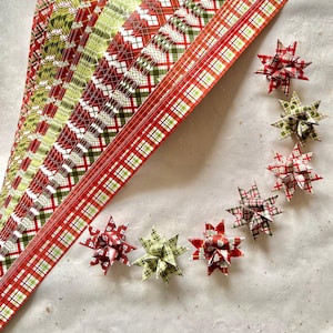 May include: A collection of red, green, and white plaid paper strips and star-shaped ornaments. The ornaments are arranged in a circle, with the paper strips fanned out in the background. The color scheme is festive.