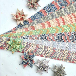 May include: Origami stars in various colors and patterns are displayed with coordinating patterned paper strips. The stars are made from paper with floral and geometric designs in shades of red, blue, green, and gray. The paper strips are arranged in a fan shape.